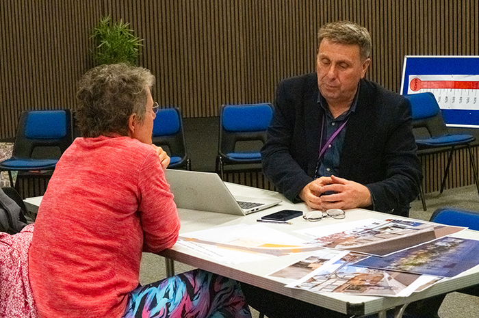 A man in a suit and a woman sit in conversation at a table covered with plans and designs